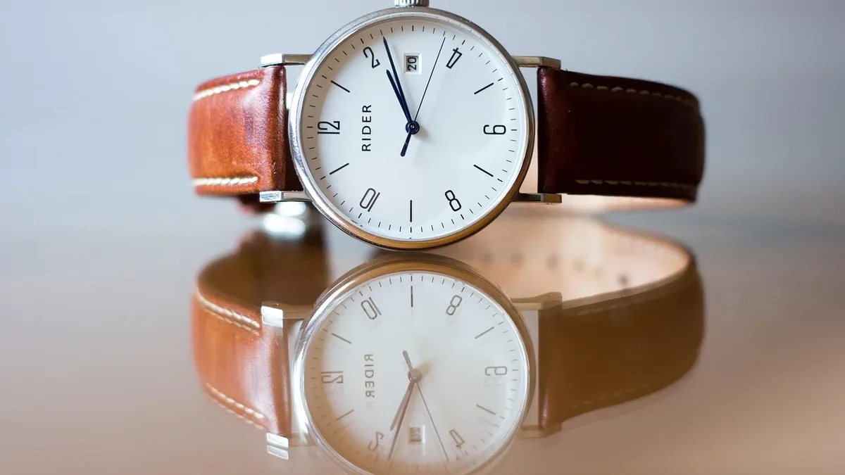 A wristwatch with a white face and brown leather strap, placed on a reflective surface.